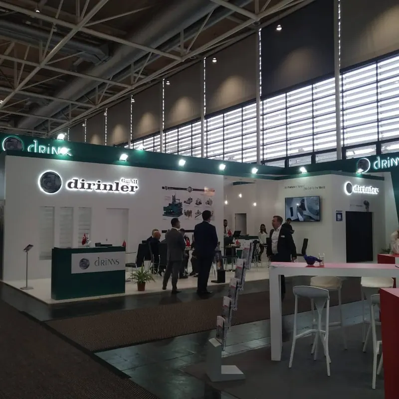 Weiß-grüner Messestand mit Maschinenexponaten, Sitzgruppen und mehreren Personen.