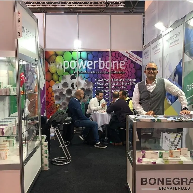 Heller Messestand mit buntem Wandbild, Glasvitrinen und besetzter Sitzgruppe.