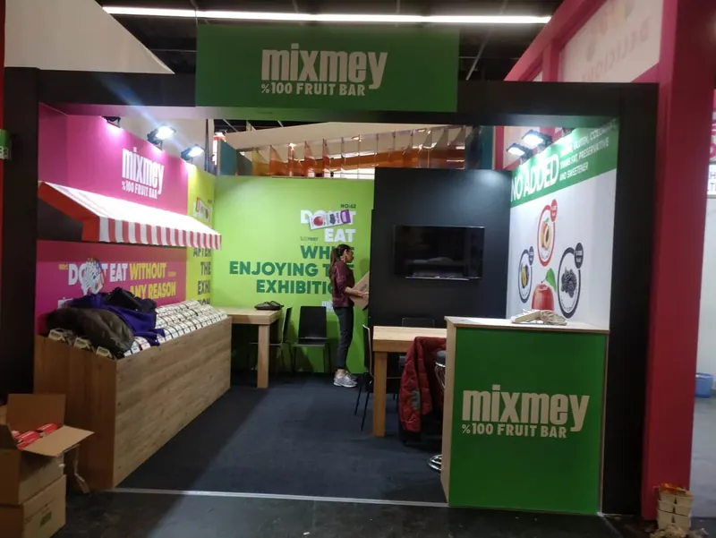 Schmaler grün-pinker Messestand mit Produktregalen, Bildschirm und Holzoptiktheke.