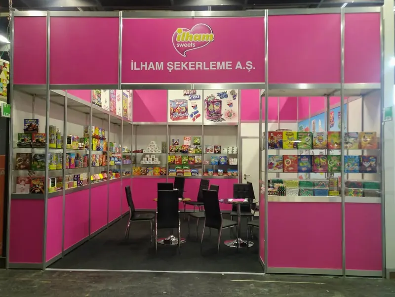 Pinker Messestand mit vielen verpackten Produkten in Vitrinen und schwarzer Sitzgruppe.