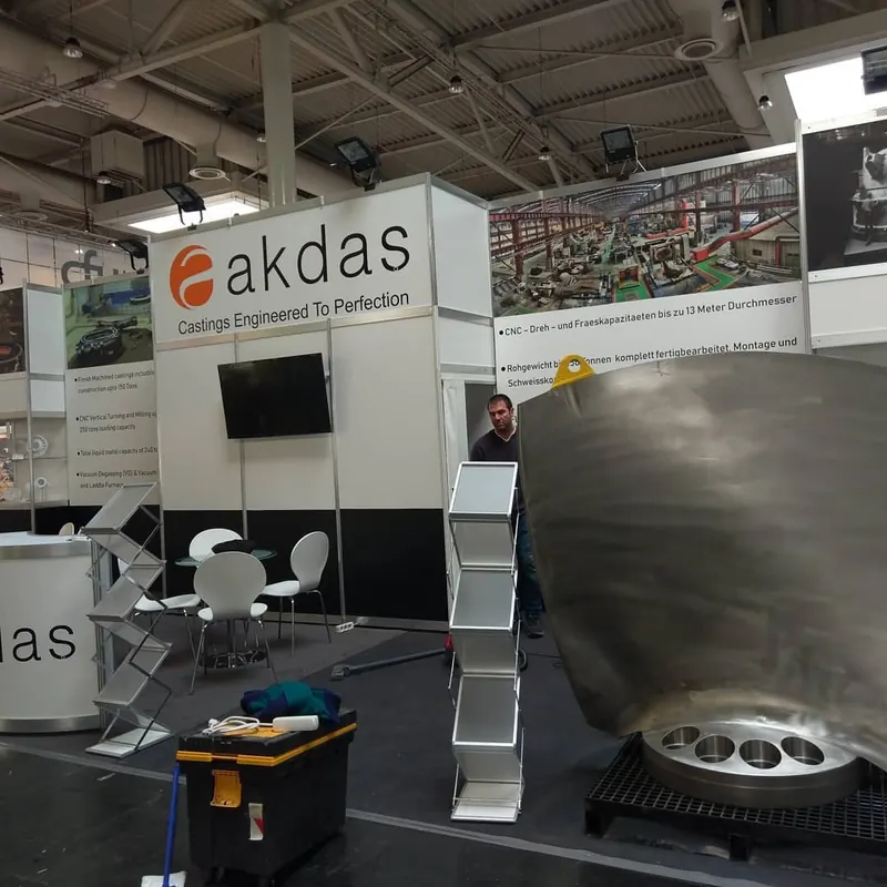 Großes gebogenes Metallteil, Prospektständer und weiße Sitzgruppe im Messestand.