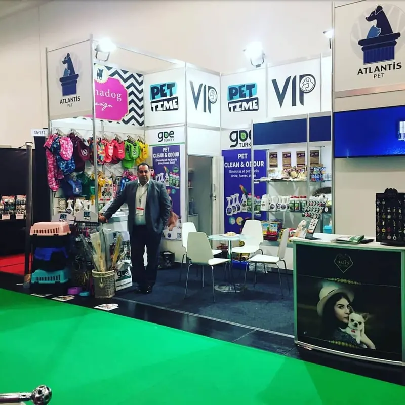 Weiß-blauer Messestand mit bunten Warenmustern, kleiner Sitzgruppe und grünem Gangteppich.