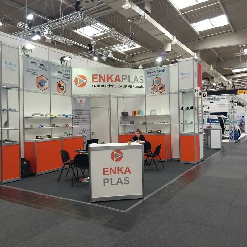 Weiß-oranger Messestand mit Produktregalen, breiter Theke und schwarzen Stühlen.