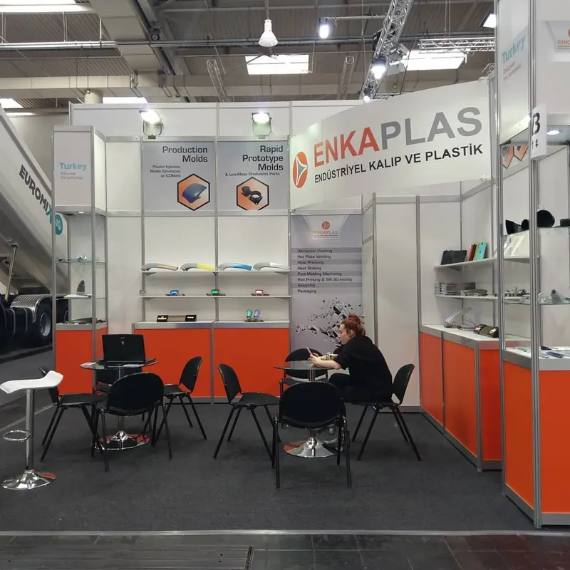 Weiß-oranger Messestand mit Glasvitrinen und schwarzen Stühlen an runden Tischen.