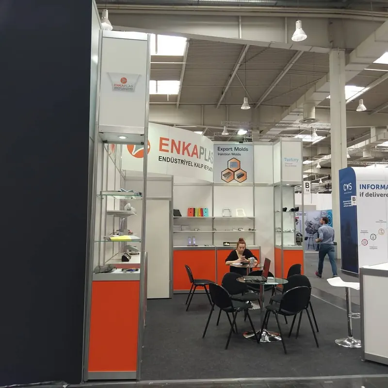 Weiß-oranger Messestand mit Glasvitrinen und schwarzen Stühlen an runden Tischen.