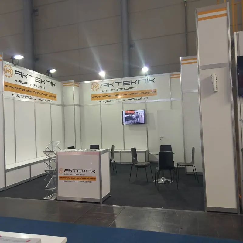 Weißer Messestand mit orangefarbenen Akzenten, Bildschirm und schwarzen Stühlen.
