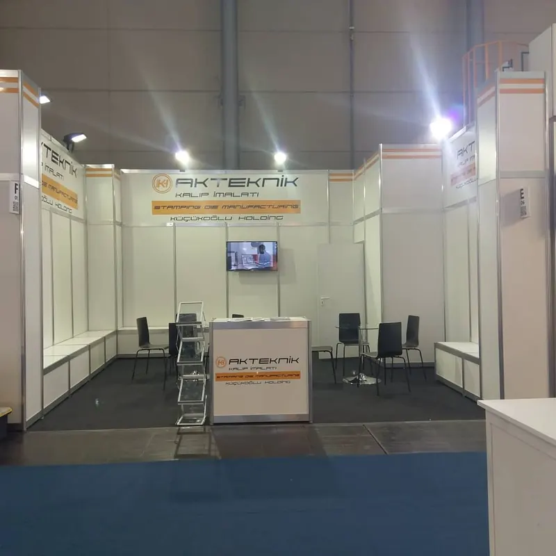 Weißer Messestand mit orangefarbenen Akzenten, Bildschirm und schwarzen Stühlen.