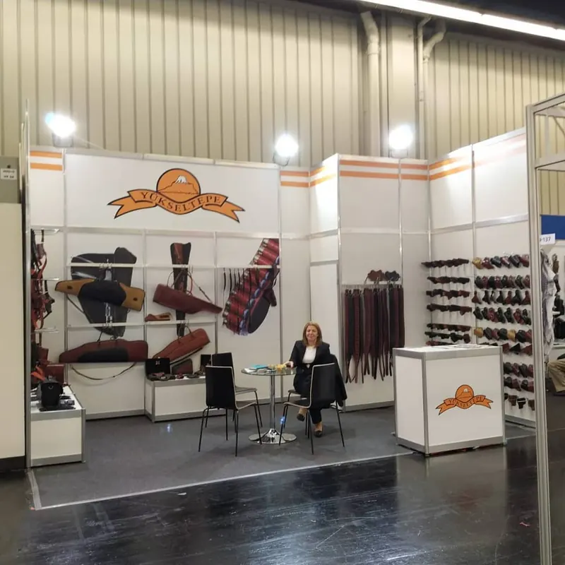 Weiß-oranger Messestand mit braunen Taschen- und Warenmustern und kleiner Sitzgruppe.