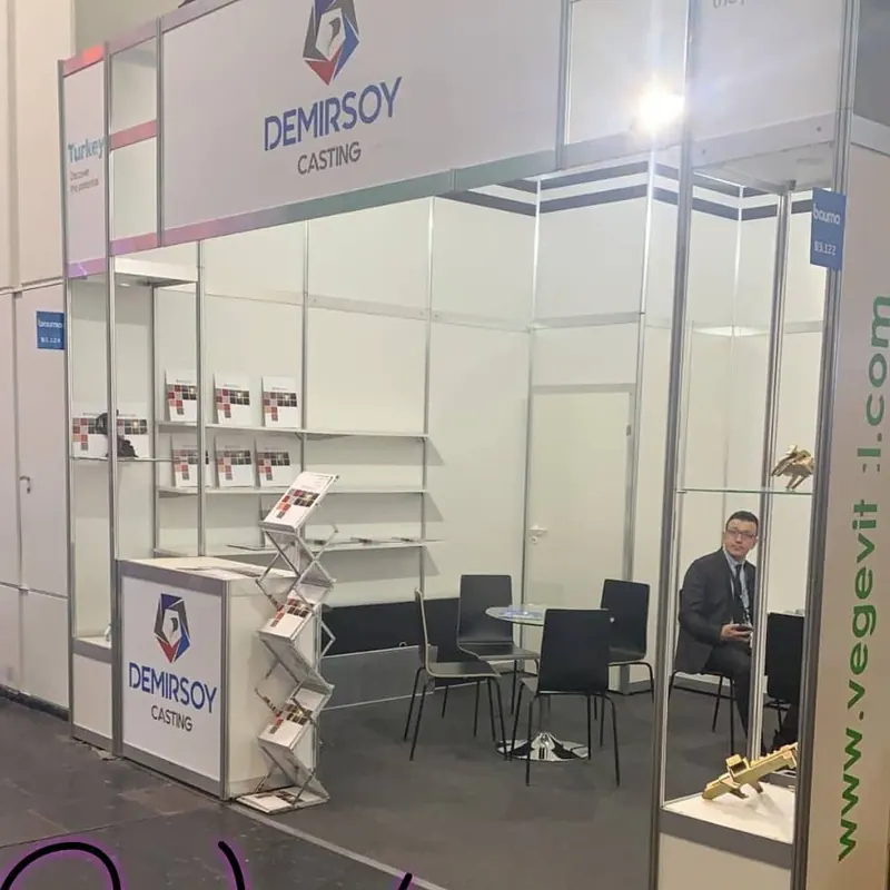 Weißer Messestand mit schmalen Regalen, Prospektständer und schwarzen Stühlen.