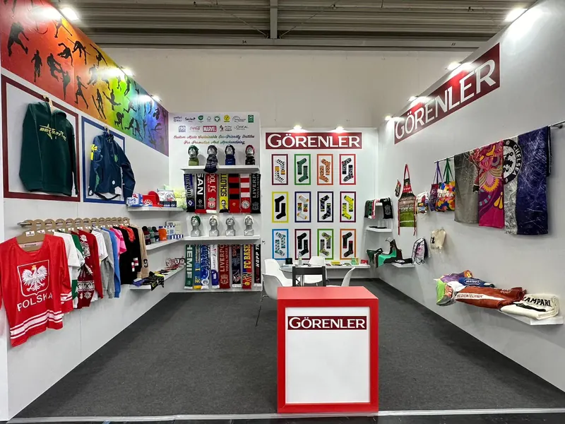 Messestand mit weißen Wänden, roter Theke und ausgestellten Trikots, Schals und Textilmustern.