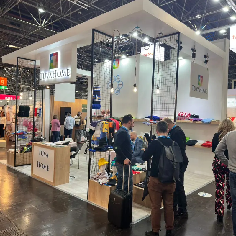 Heller Messestand mit dunklen Gitterstützen, Stoffmustern und Besuchern im Gang.