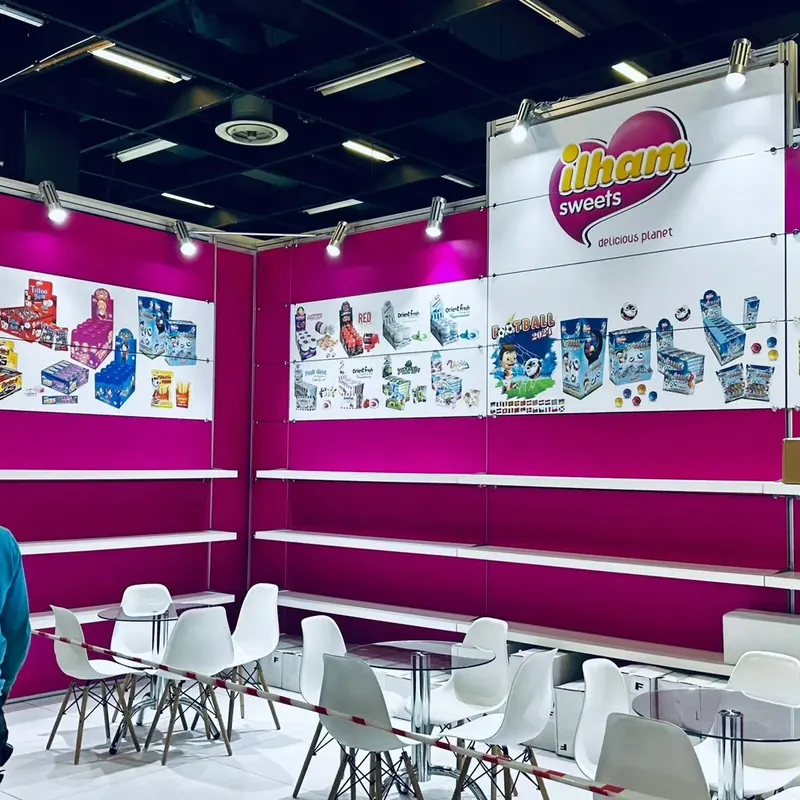 Magenta-weißer Messestand mit langen Wandregalen und mehreren weißen Tischgruppen.