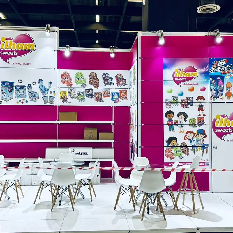 Pink-weißer Messestand mit bunten Verpackungsmotiven, Regalen und weißen Sitzgruppen.