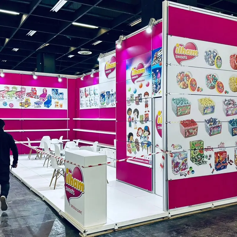 Pink-weißer Messestand mit bunten Verpackungsmotiven, Regalen und weißen Sitzgruppen.