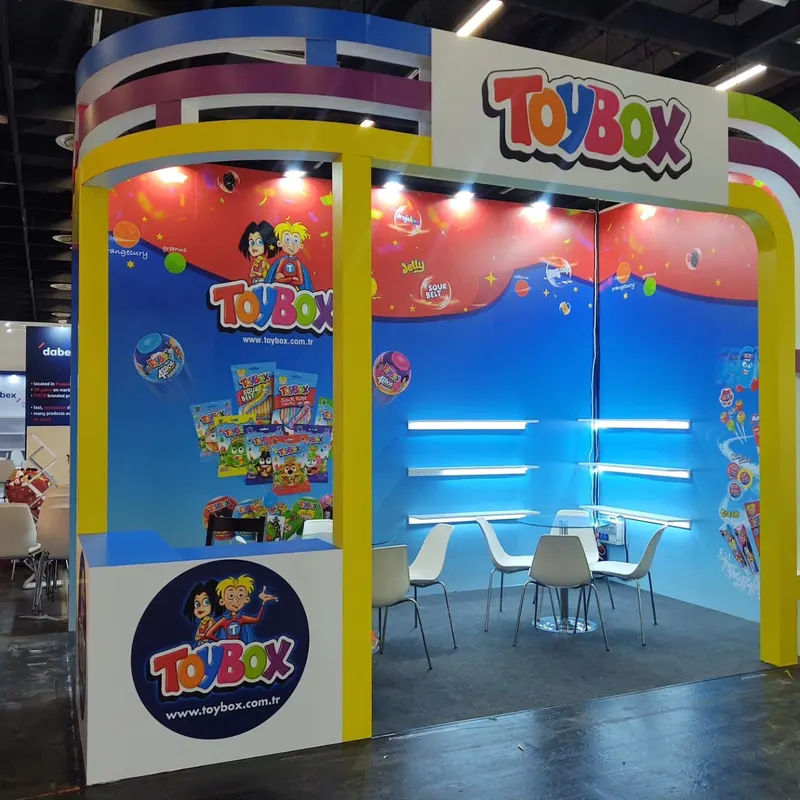 Bunter Messestand mit gelbem Rahmen, blauen Wänden, Figurenmotiven und weißen Sitzgruppen.