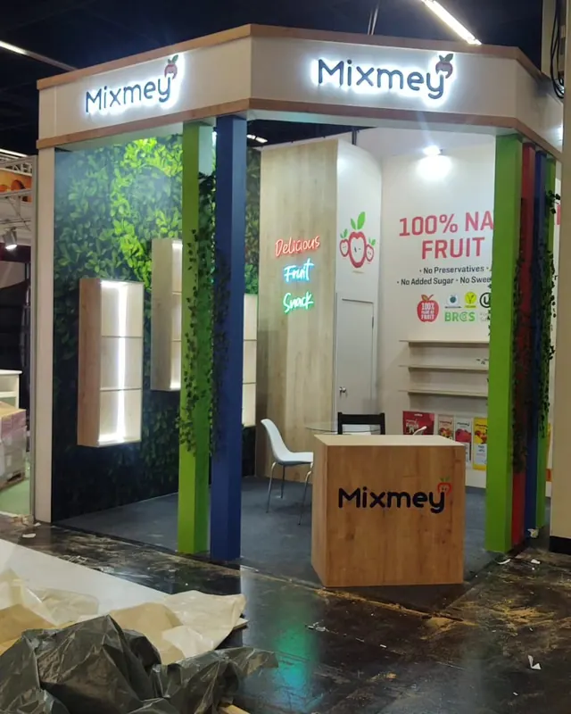 Heller Messestand mit grünen Stützen, Fruchtgrafiken und Holzoptiktheke.