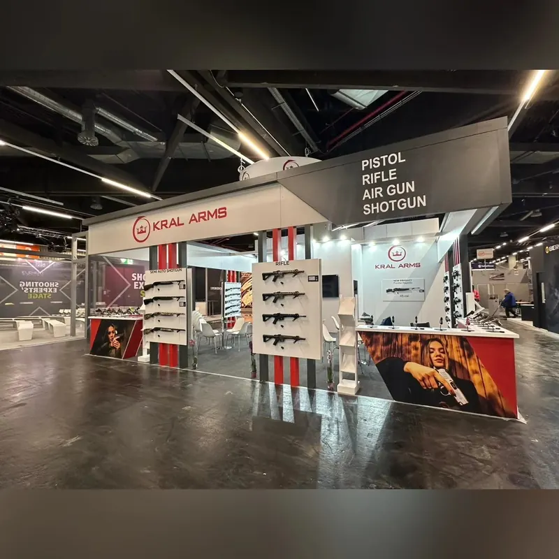 Weiß-grauer Messestand mit abgehängtem Rahmen, Langwaffen an Wanddisplays und Bildtheke.