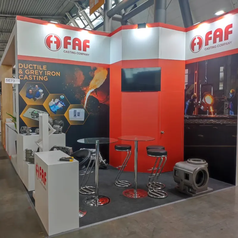 Rot-weißer Messestand mit großen Metallteilen auf Podesten und schwarzen Barhockern.