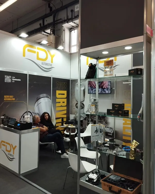 Schwarz-gelber Messestand mit Produktvitrinen, Sitzgruppe und Personen am Tisch.