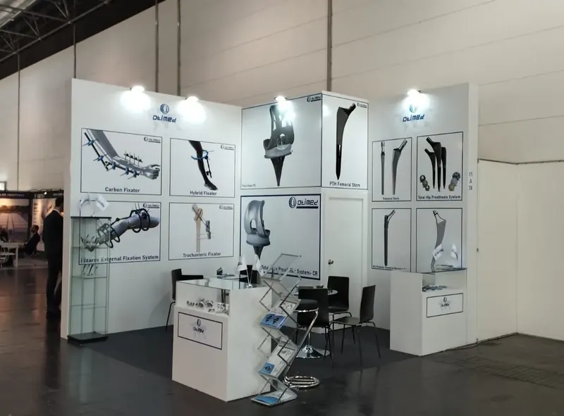 Weißer Messestand mit großen Bauteilzeichnungen, kleiner Theke und schwarzen Stühlen.