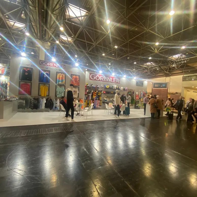 Weißer Messestand mit bunten Textilien und mehreren Personen am Hallengang.