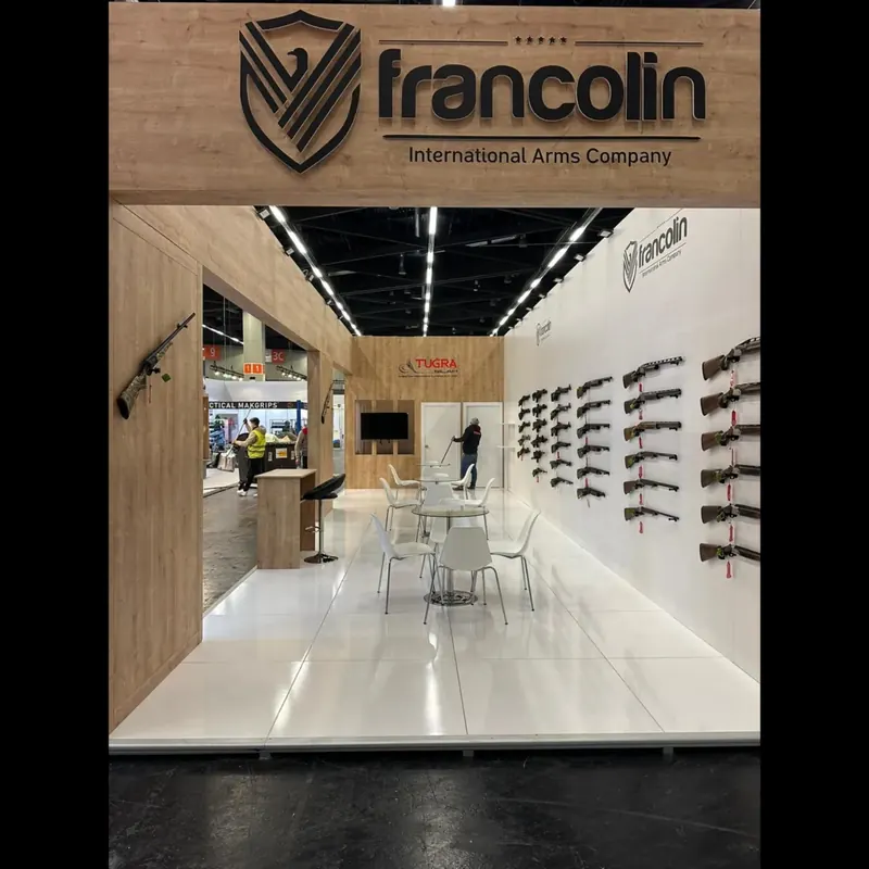 Blick durch einen Holzrahmen auf einen weißen Messestand mit Langwaffen an der Wand.