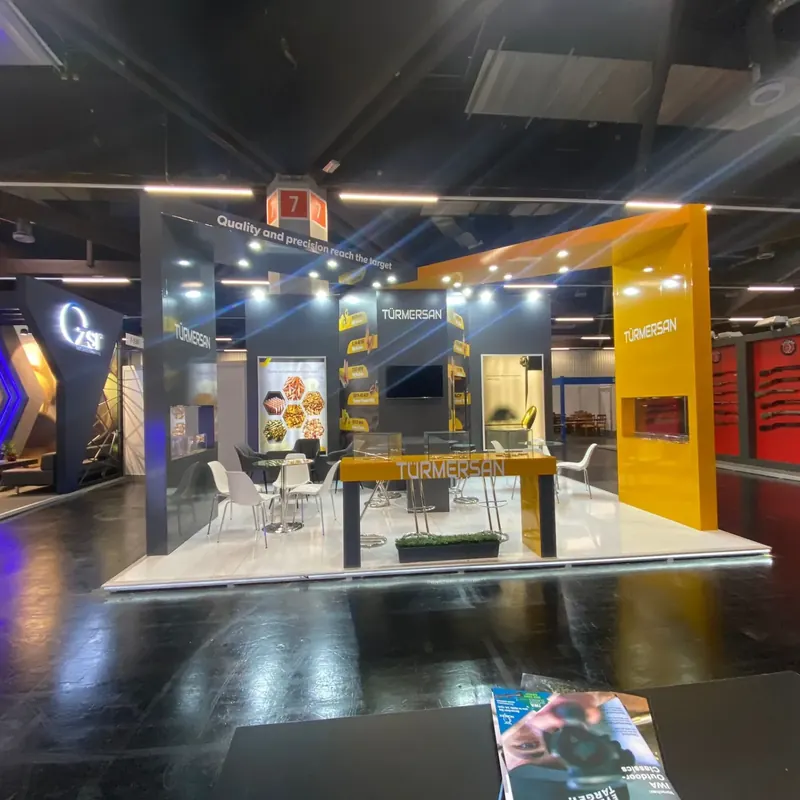 Dunkler Messestand mit gelbem Eckportal, hellem Boden und kleiner Sitzgruppe.