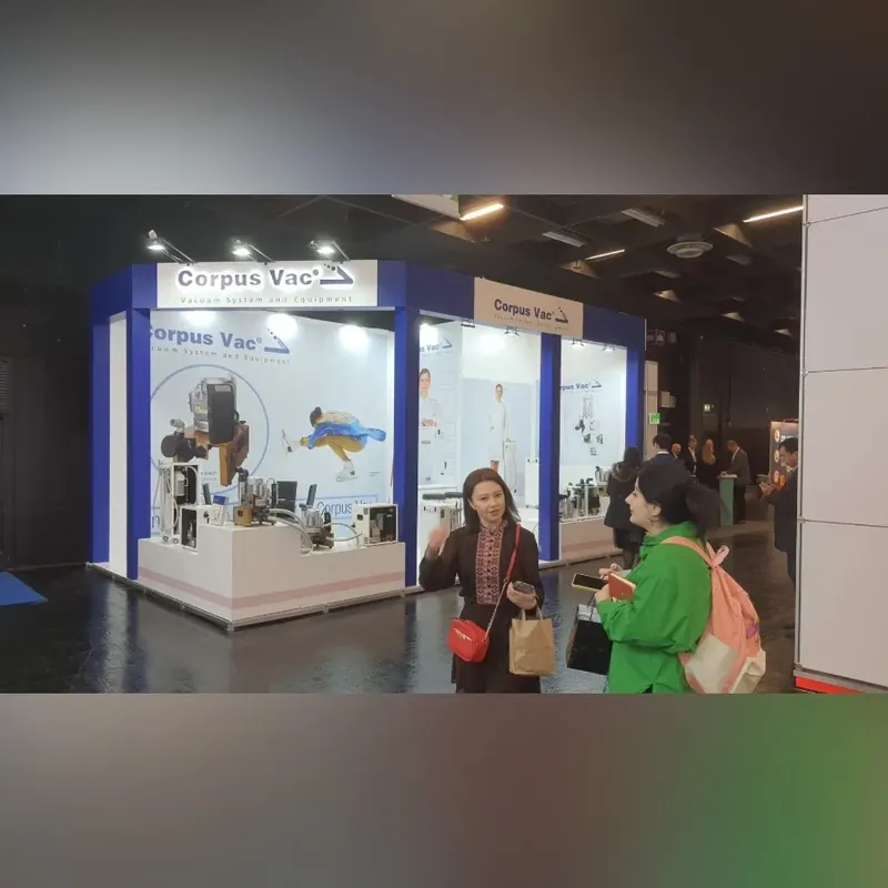 Zwei Personen im Hallengang vor einem weiß-blauen Messestand mit Produktpodesten.