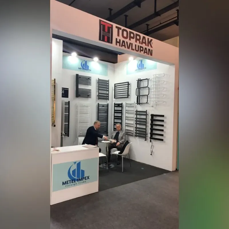 Weißer Messestand mit Wandmustern verschiedener Heizkörper und kleiner Sitzgruppe.