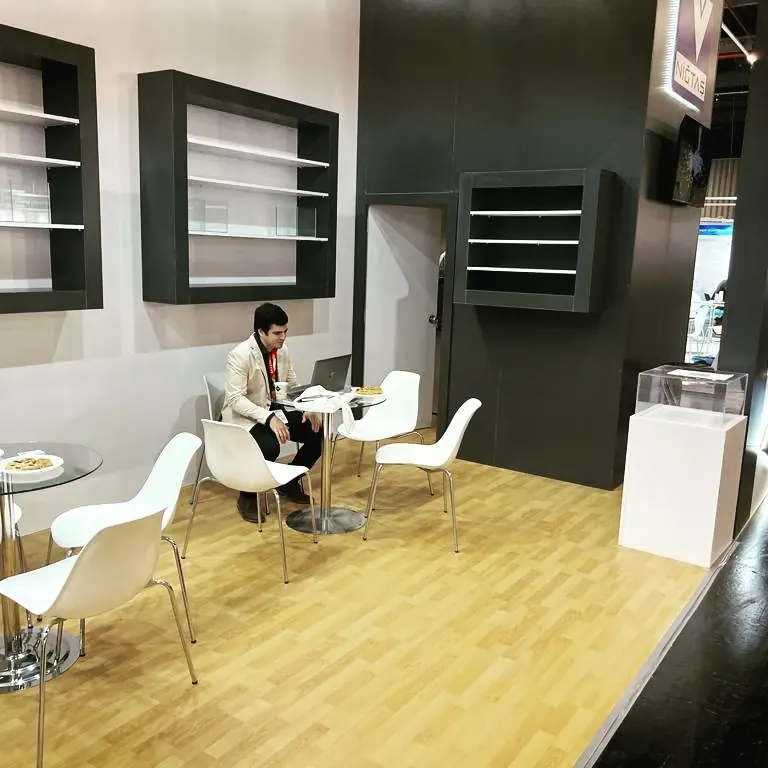 Weiß-schwarzer Messestand mit Wandvitrinen, Holzboden und einer Person an der Sitzgruppe.