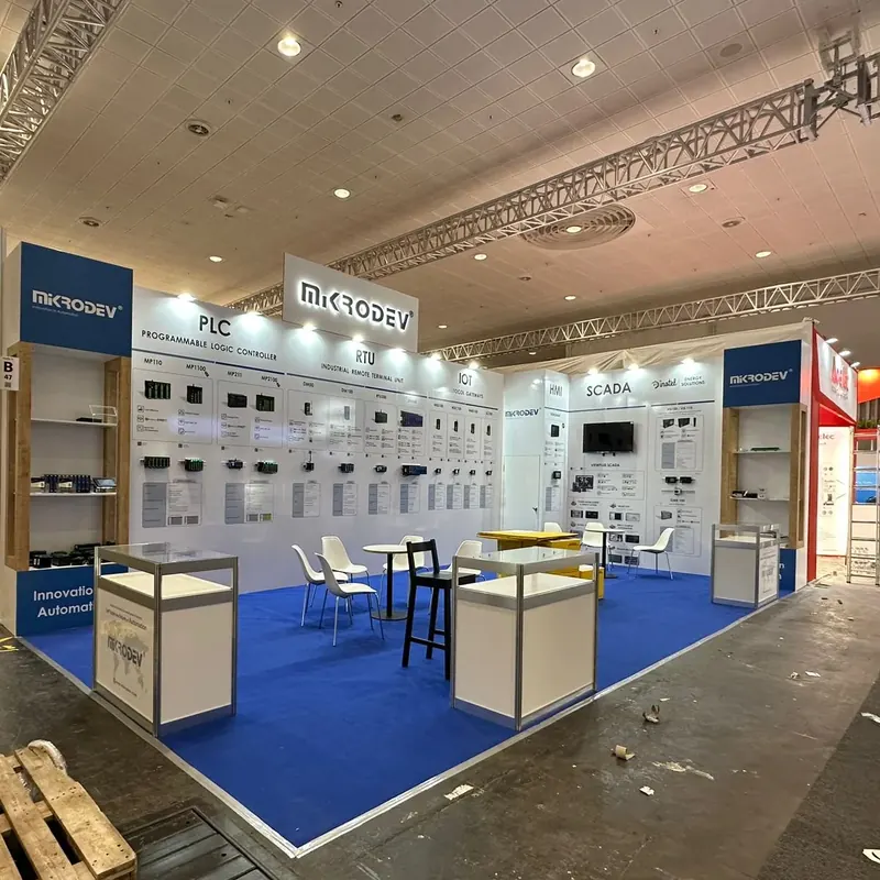 Weiß-blauer Messestand mit technischen Wandmustern, Glasvitrinen und kleiner Sitzgruppe.