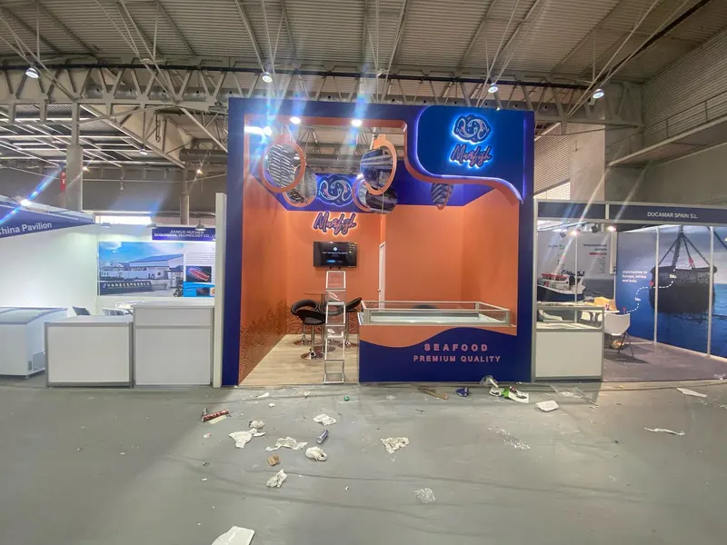 Orange-blauer Messestand mit hohem Mittelaufbau, Produktvitrinen und Bildschirmen.