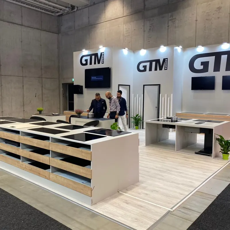 Weißer Messestand mit langen Arbeitsplatten, schwarzen Spülbecken und Personen an der Theke.