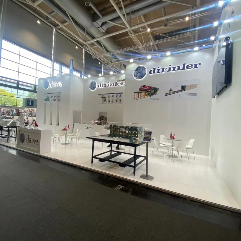 Weißer Messestand mit Maschinenteilen auf schwarzen Tischen und weißen Sitzgruppen.