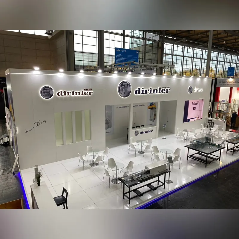 Weißer Messestand mit Sitzgruppen, schwarzen Maschinentischen und blau beleuchteter Bodenkante.