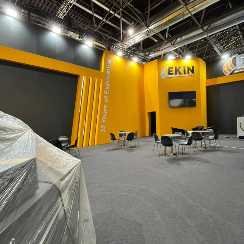 Gelb-dunkelgrauer Messestand mit schwarzen Stühlen und abgedecktem Exponat im Vordergrund.