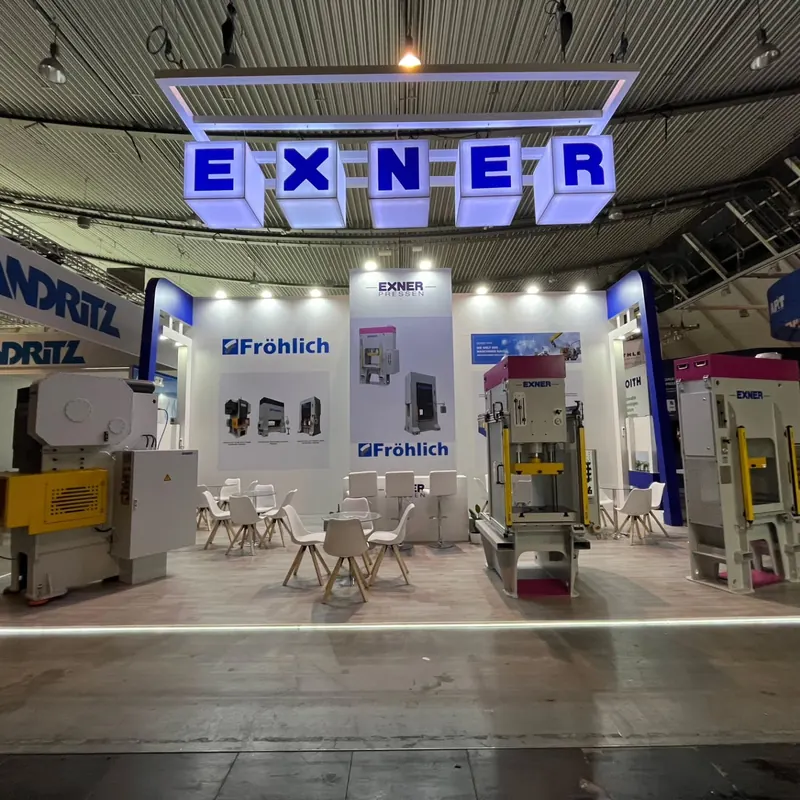 Weiß-blauer Messestand mit beleuchteten Buchstabenwürfeln, Maschinen und Sitzgruppe.