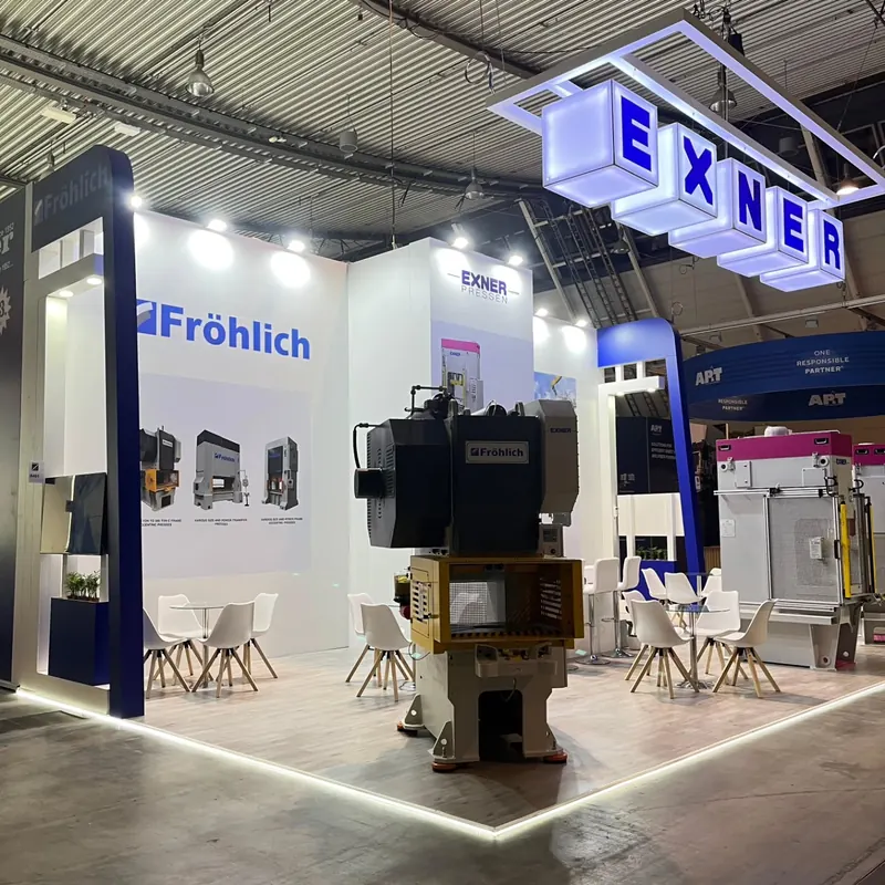 Weiß-blauer Messestand mit beleuchteten Buchstabenwürfeln, Maschinen und Sitzgruppe.