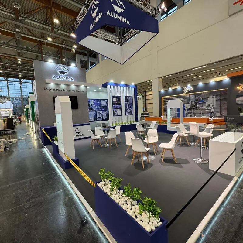 Blau-weißer Messestand mit abgehängtem Würfelschild, Produktgrafiken und weißen Stühlen.