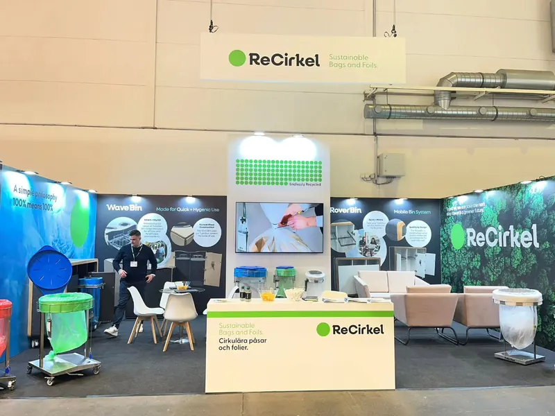 Messestand mit grün-weißer Rückwand, Produktbehältern, Sitzgelegenheiten und einer Theke.
