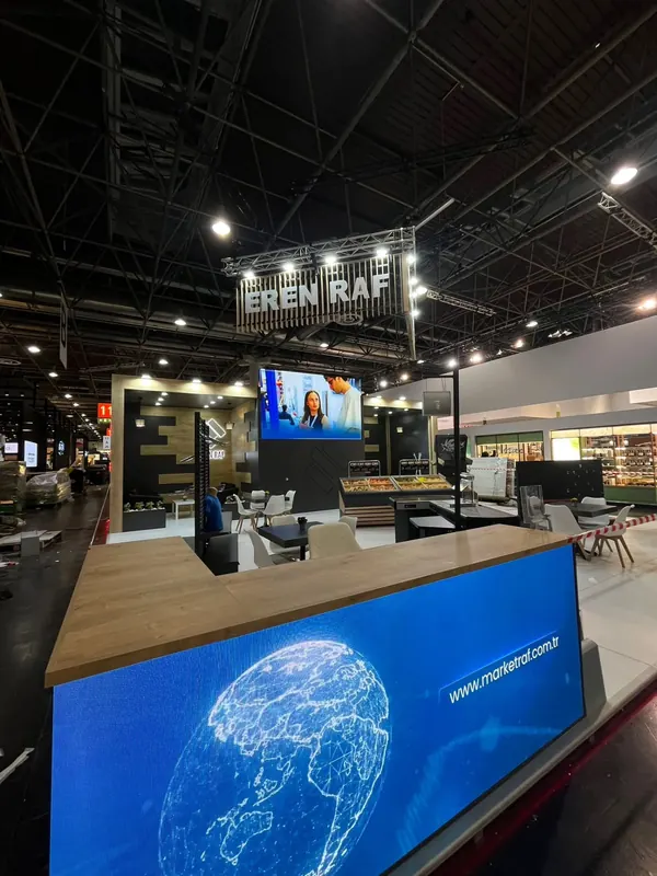 Dunkler Messestand mit Holzoptiktheke, großem blauem Leuchtbild und Bildschirmen.
