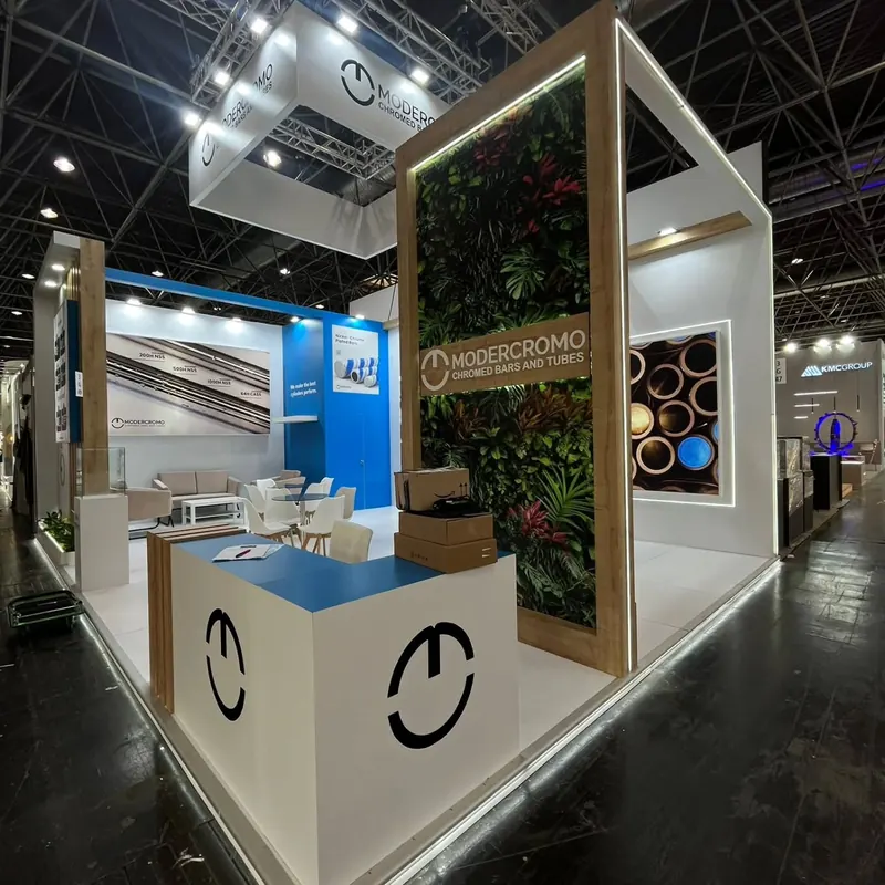 Weißer Messestand mit hoher Pflanzenwand, Holzoptikrahmen und blauer Empfangstheke.