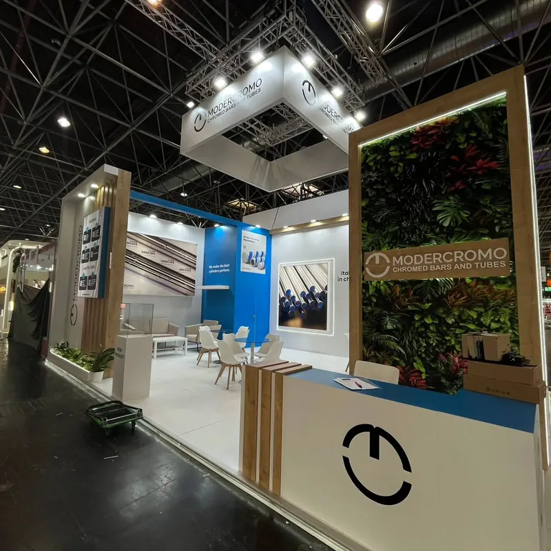 Weißer Messestand mit Holzlamellen, grüner Pflanzenwand und blau-weißer Empfangstheke.