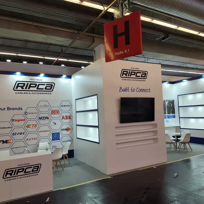 Weißer Messestand mit beleuchteten Wandregalen, dunklem Bildschirm und kleiner Theke.